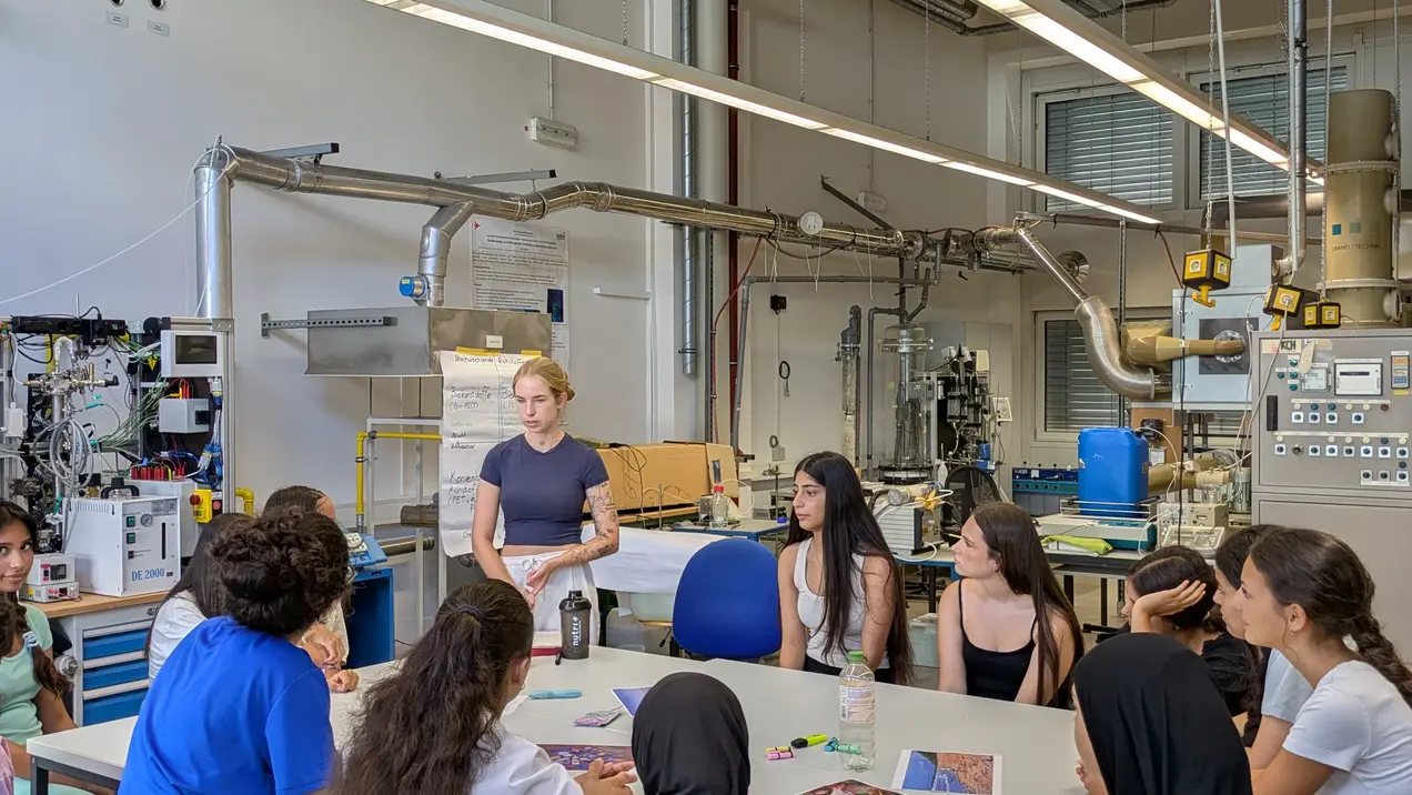 Eine Lehrerin steht vor einer Gruppe von Schülerinnen und Schülern in einem modernen Labor. Die Teilnehmer sitzen an einem Tisch und betrachten Materialien, während sie aufmerksam zuhören. Im Hintergrund sind technische Geräte sichtbar, die für den Unterricht verwendet werden.