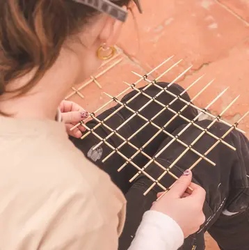 Eine Person sitzt mit einer Struktur aus Holzstäben auf dem Schoß. Sie hält die Stäbe in der Hand und arbeitet daran, während sie von hinten fotografiert wird. Die Umgebung besteht aus einem roten, ungepflegten Boden.