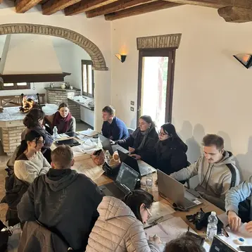 A group of about 15 architectural heritage students sit at a long table in a bright room with a wooden ceiling. They are all busy on their laptops, some taking notes. A few conversations seem to be taking place as light streams in through the windows. They are on an excursion in Italy.