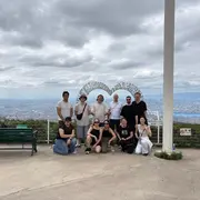 Eine Gruppe von zehn Personen steht in einer hügeligen Landschaft. Im Hintergrund ist eine Stadtlandschaft zu sehen, und ein großes, herzförmiges Kunstwerk ist sichtbar. Die Personen lächeln und posieren für das Foto, während der Himmel teilweise bewölkt ist.