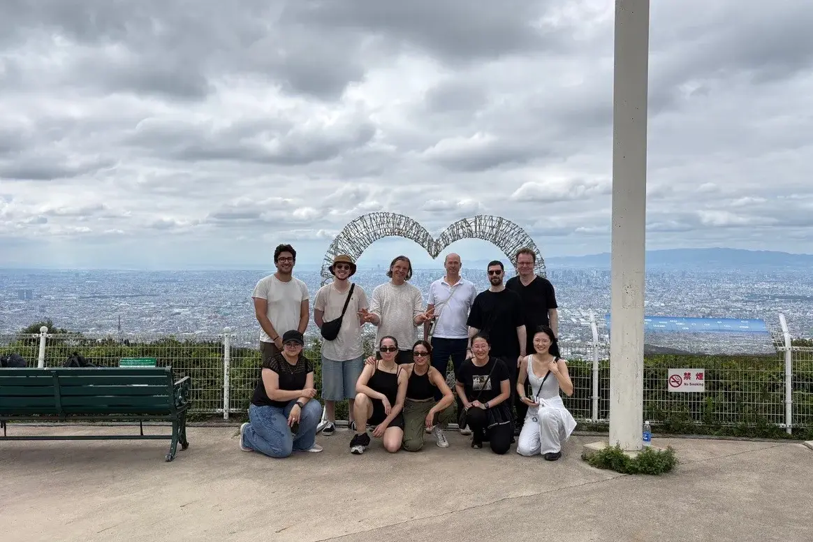 Eine Gruppe von zehn Personen steht in einer hügeligen Landschaft. Im Hintergrund ist eine Stadtlandschaft zu sehen, und ein großes, herzförmiges Kunstwerk ist sichtbar. Die Personen lächeln und posieren für das Foto, während der Himmel teilweise bewölkt ist.