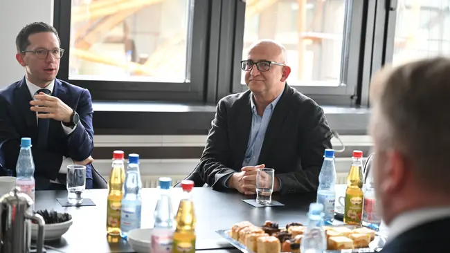 Zwei Männer sitzen an einem Tisch in einem heller eingerichteten Raum. Der Mann links trägt einen Anzug und spricht. Der Mann rechts trägt eine Brille und hört aufmerksam zu. Auf dem Tisch befinden sich Getränke in Flaschen und Teller mit Süßigkeiten.