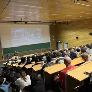 Ein Raum mit vielen Menschen, die einer Präsentation lauschen. Vorne stehen zwei Referenten, während auf einer großen Leinwand Informationen zu sehen sind. Die Atmosphäre ist konzentriert und die Zuhörer sitzen in mehreren Reihen an Tischen.