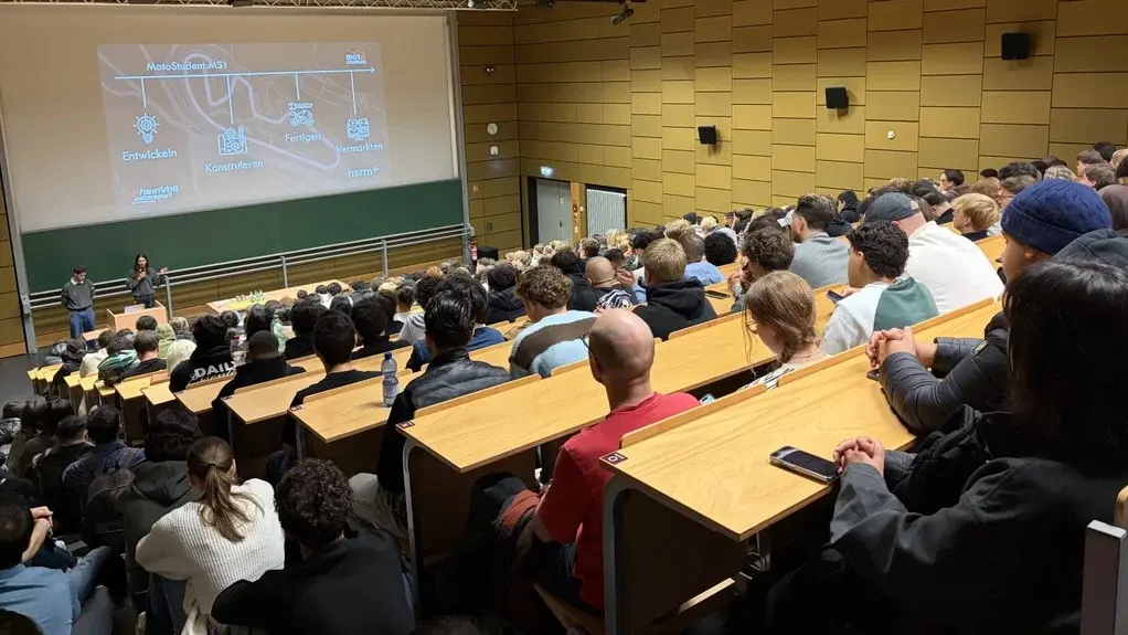 Ein Raum mit vielen Menschen, die einer Präsentation lauschen. Vorne stehen zwei Referenten, während auf einer großen Leinwand Informationen zu sehen sind. Die Atmosphäre ist konzentriert und die Zuhörer sitzen in mehreren Reihen an Tischen.