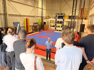 Eine Gruppe von Menschen steht vor einem Trainingsbereich mit einer blauen Matte und bunten Turngeräten. Ein Mann erklärt etwas, während die Zuhörer aufmerksam zuhören. Der Raum ist geräumig und gut beleuchtet, mit Sicherheitsnetzen und Turngeräten im Hintergrund.