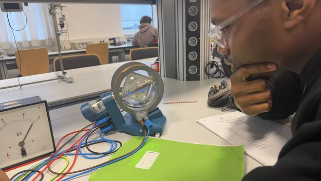 Ein Schüler sitzt an einem Tisch in einem Labor und betrachtet ein Experiment. Auf dem Tisch befinden sich ein Messgerät, ein Motor und verschiedene Kabel. Der Mann scheint tief in Gedanken versunken, während er sich auf die Analyse der Ergebnisse konzentriert.