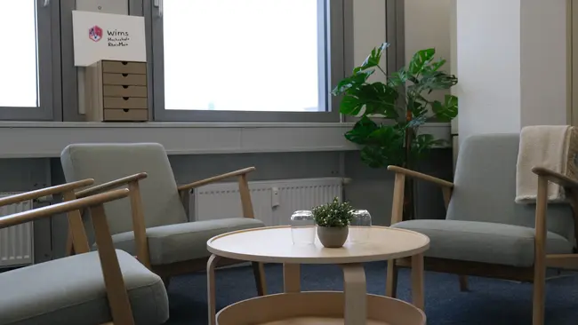 In einem hellen Raum befinden sich zwei graue Sessel und ein runder Holztisch mit einer kleinen Pflanze in der Mitte. Im Hintergrund sind Fenster und eine grüne Pflanze sichtbar. An der Wand hängt eine Box mit dem Logo „wirms“.