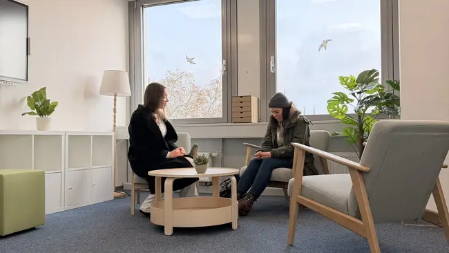 Zwei Frauen sitzen in einem hellen Raum mit großen Fenstern. Eine Frau mit langen Haaren trägt einen schwarzen Mantel und spricht, während die andere in einem grauen Pullover und Mütze auf ihr Handy schaut. Zwischen ihnen steht ein runder Tisch mit einer kleinen Pflanze.