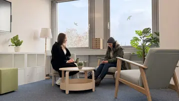 Zwei Frauen sitzen in einem hellen Raum mit großen Fenstern. Eine Frau mit langen Haaren trägt einen schwarzen Mantel und spricht, während die andere in einem grauen Pullover und Mütze auf ihr Handy schaut. Zwischen ihnen steht ein runder Tisch mit einer kleinen Pflanze.