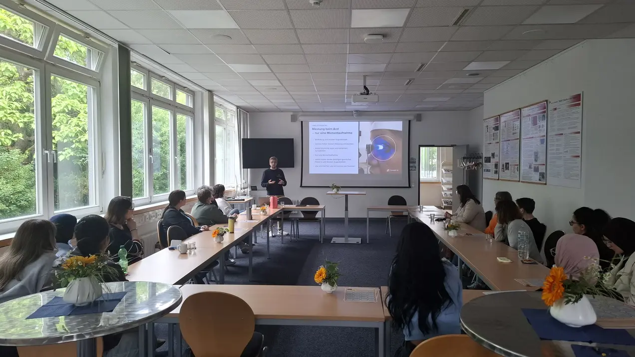 Eine Person steht vorne in einem Konferenzraum und präsentiert an einer Wand, wo ein Bildschirm mit Diagrammen zu sehen ist. Im Raum sitzen etwa 15 Zuhörer, die aufmerksam lauschen. Es gibt dekorierte Tische mit Blumen. Tageslicht strömt durch große Fenster.