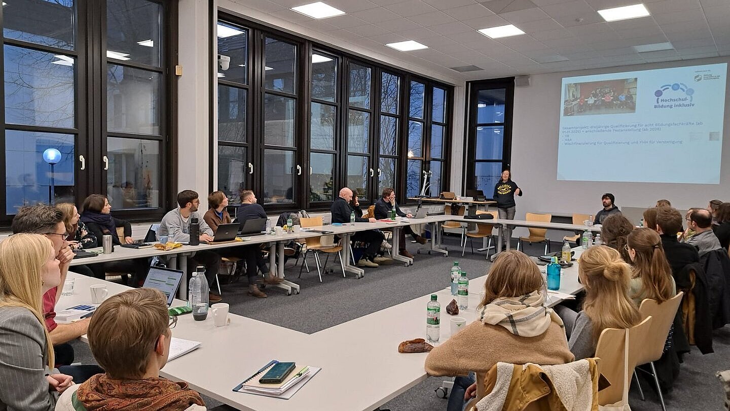 Tagung. Vortrag vor einer Gruppe Menschen, die im Kreis an Tischen sitzen