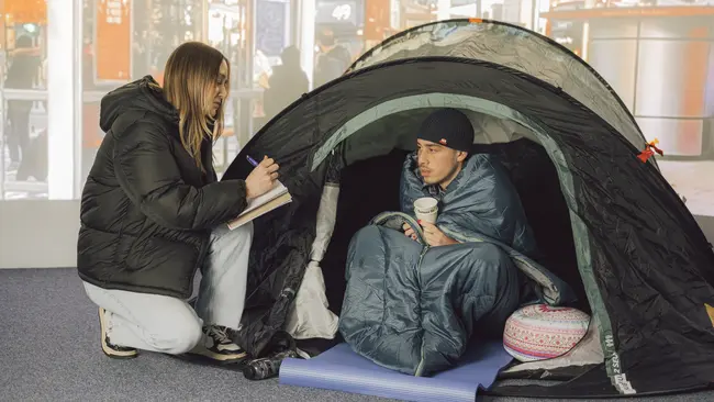 Zwei Studies, die eine Obdachlosenszene darstellen, wo eine Person im Zelt, eingewickelt in einem Schlafsack sitzt und traurig zum Boden schaut. Die andere Person kniet sich zu ihr nieder und schreibt sich die Situation auf. Im Hintergrund ist eine Bahnhofsszene abgebildet.