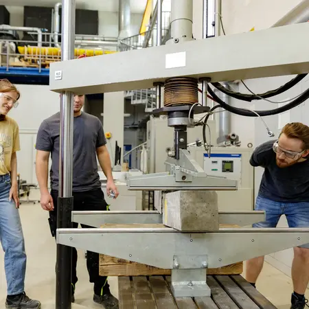 Eine Gruppe von vier Studierenden beobachtet einen Experimentieraufbau im Labor für Massivbau. Im Mittelpunkt steht eine pressende Maschine, die ein Betonteil testet.