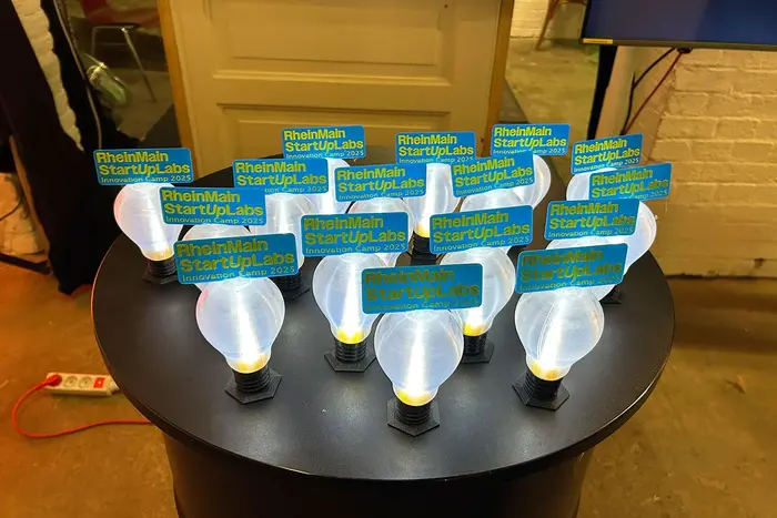 Auf einem Tisch stehen kleine, mit dem 3-D-Drucker erstellte Glühbirnen, auf denen der Schriftzug "RheinMain StartUpLabs | Innovation Camp 2025 zu lesen ist.