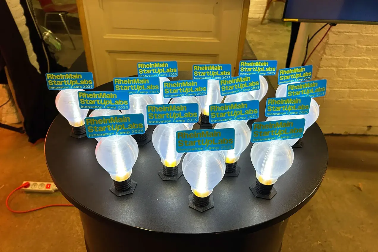 Auf einem Tisch stehen kleine, mit dem 3-D-Drucker erstellte Glühbirnen, auf denen der Schriftzug "RheinMain StartUpLabs | Innovation Camp 2025 zu lesen ist.