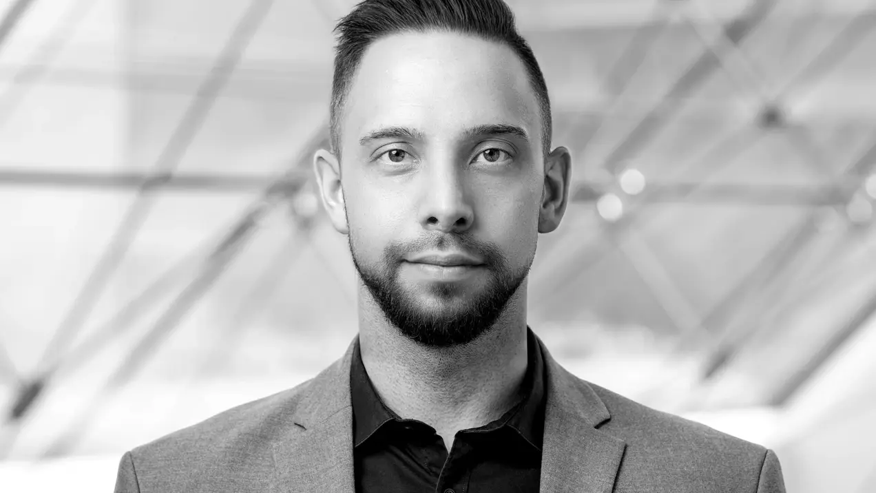 Porträt eines Mannes in einem eleganten grauen Anzug mit schwarzem Hemd. Er blickt direkt in die Kamera, hat eine neutrale Miene und trägt gepflegte Haare. Der Hintergrund ist unscharf und zeigt geometrische Linien, die ein modernes Umfeld andeuten.