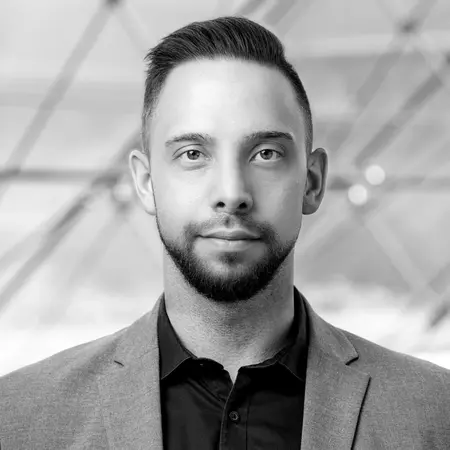 Porträt eines Mannes in einem eleganten grauen Anzug mit schwarzem Hemd. Er blickt direkt in die Kamera, hat eine neutrale Miene und trägt gepflegte Haare. Der Hintergrund ist unscharf und zeigt geometrische Linien, die ein modernes Umfeld andeuten.