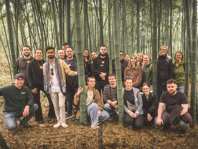 Eine Gruppe von 24 Personen steht in einem Bambuswald. Sie lächeln und posieren zusammen, während sie zwischen den hohen Bambusstämmen arrangiert sind. Die Atmosphäre ist freundlich und entspannt.
