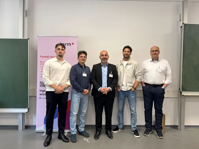 Fünf Personen stehen nebeneinander vor einer Wand. Im Hintergrund befindet sich ein Banner mit dem Logo eines Unternehmens. Die Personen sind in formeller Kleidung und lächeln. Die Umgebung wirkt wie ein Klassenzimmer oder eine Präsentationsfläche.