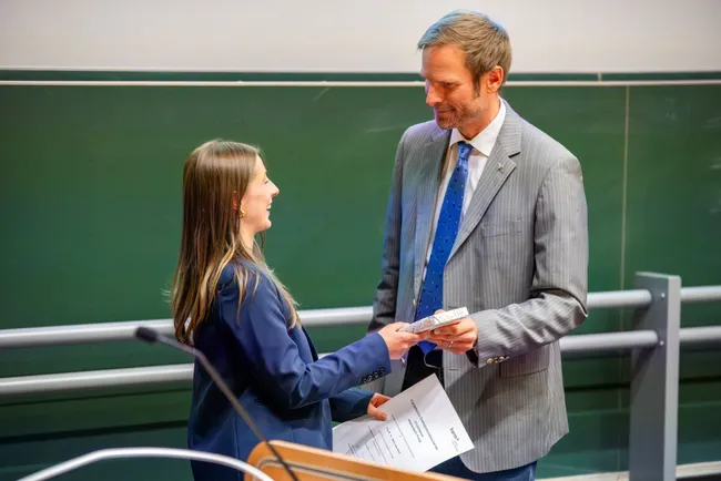 Eine Frau in einem blauen Blazer überreicht ein Dokument an einen Mann in einem grauen Anzug während einer Zeremonie. Beide lächeln sich an. Im Hintergrund ist eine Tafel zu sehen.