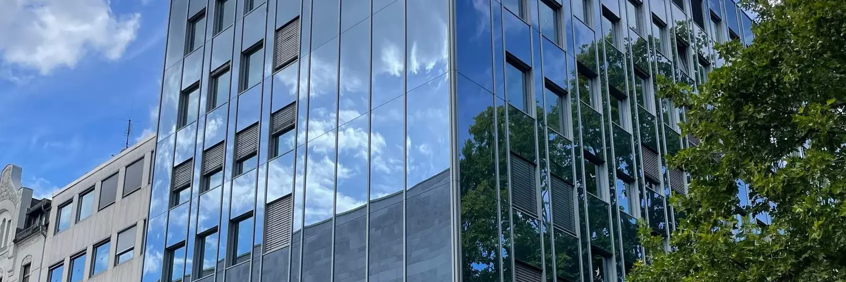 Ein moderner, mehrstöckiger Bürogebäude mit einer Glasfassade, die den Himmel und die umgebenden Bäume reflektiert. Vor dem Gebäude sind einige Sitzgelegenheiten und eine breite Straße. Der Himmel ist teils bewölkt.