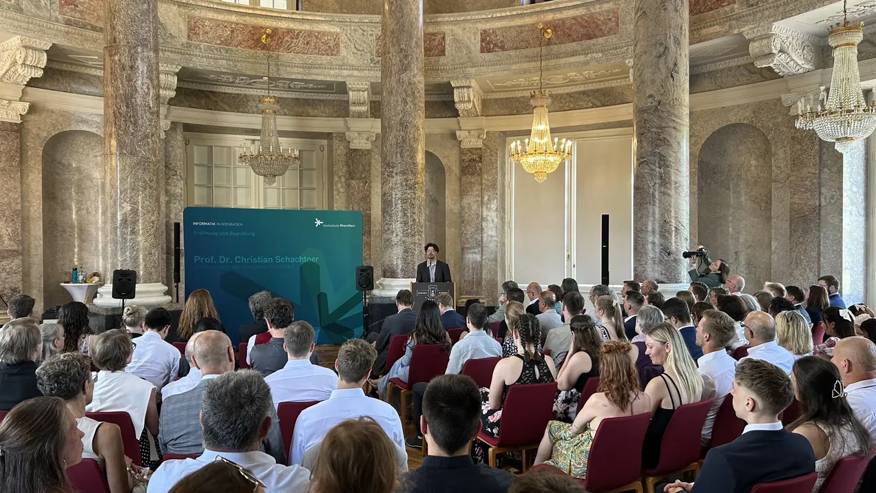 Eine Menschenmenge sitzt in einem eleganten Raum mit hohen Decken und Säulen. Im Vordergrund steht ein Redner am Pult und hält eine Ansprache. Im Hintergrund sind einige Zuhörer zu sehen, während andere aufmerksam in die Runde blicken.