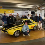 Gruppenfoto von Menschen vor einem gelben Oldtimer-Auto in einer modernen Ausstellung. Die Personen tragen Ansteckschilder und lächeln, während sie sich um das Fahrzeug gruppieren. Im Hintergrund sind weitere Autos und ein Ausstellungsbereich sichtbar.