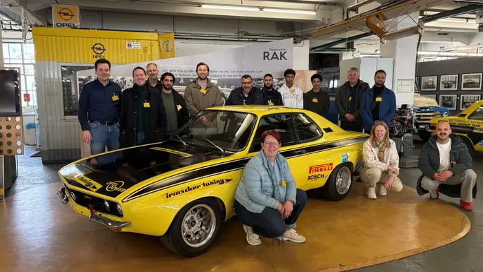 Gruppenfoto von Menschen vor einem gelben Oldtimer-Auto in einer modernen Ausstellung. Die Personen tragen Ansteckschilder und lächeln, während sie sich um das Fahrzeug gruppieren. Im Hintergrund sind weitere Autos und ein Ausstellungsbereich sichtbar.