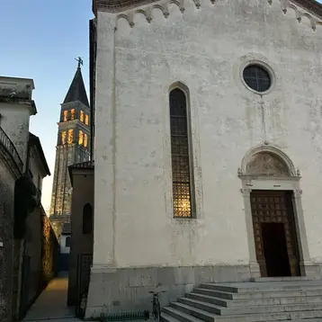 A historic building with a light-colored facade and large windows, flanked by a staircase with several steps. In the background, a tall church tower rises, its windows shining in the light, while the sky glows at dusk.