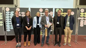 Sechs Teilnehmende der Tagung posieren für ein Gruppenbild. Unter ihnen der diesjährige Gastgeber Prof. Dr.-Ing. Michael Kloos von der HSRM. Außerdem die ehemaligen sowie neu gewählten Vorsitzenden der UNESCO Lehrstühle in Deutschland. Der verabschiedete Vorsitzende hält ein Geschenk in den Händen.