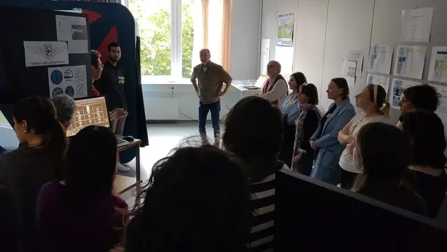 Eine Gruppe von Menschen steht in einem Raum und hört einem Vortrag zu. Im Vordergrund spricht eine Person, während eine andere Person hinter einem Tisch mit Informationen steht. Im Hintergrund sind Dokumente und Plakate an den Wänden sichtbar. Licht fällt durch ein Fenster.