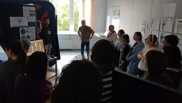 Eine Gruppe von Menschen steht in einem Raum und hört einem Vortrag zu. Im Vordergrund spricht eine Person, während eine andere Person hinter einem Tisch mit Informationen steht. Im Hintergrund sind Dokumente und Plakate an den Wänden sichtbar. Licht fällt durch ein Fenster.