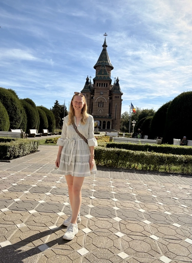 Eine junge Frau in einem hellen Kleid steht auf einem gepflasterten Weg in einem Park. Im Hintergrund ist ein historisches Gebäude mit Turm und architektonischen Details sichtbar. Der Himmel ist blau mit einigen Wolken, und das Gelände ist von gepflegten Hecken umgeben.
