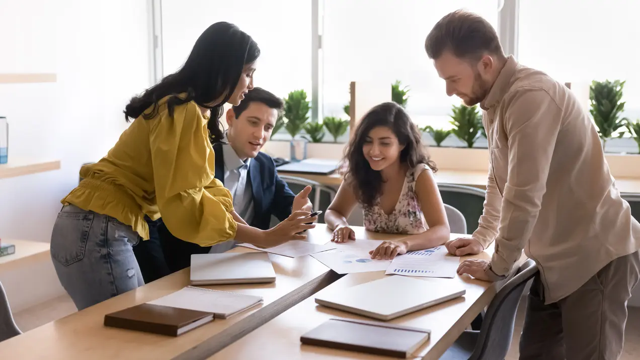 Vier Personen, zwei Männer und zwei Frauen, stehen um einen Tisch und diskutieren über Dokumente. Eine Frau zeigt etwas auf ihrem Handy, während die anderen aufmerksam schauen. Der Raum ist hell und modern eingerichtet, mit Pflanzen im Hintergrund.