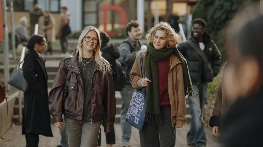 Zwei junge Frauen gehen lächelnd nebeneinander auf einem belebten Weg. Eine trägt eine braune Lederjacke und die andere einen dicken Schal mit einer großen Tasche. Im Hintergrund sind weitere Personen, die an einem warmen Tag draußen unterwegs sind.