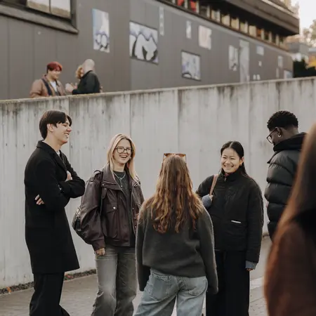 Sechs Personen stehen in einer Gruppe und unterhalten sich lebhaft. Sie tragen lässige Kleidung und wirken freundlich. Im Hintergrund sind Gebäude und Wandkunst zu sehen, während einige Personen verstreut im Hintergrund stehen.
