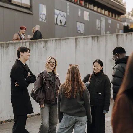 Sechs Personen stehen in einer Gruppe und unterhalten sich lebhaft. Sie tragen lässige Kleidung und wirken freundlich. Im Hintergrund sind Gebäude und Wandkunst zu sehen, während einige Personen verstreut im Hintergrund stehen.