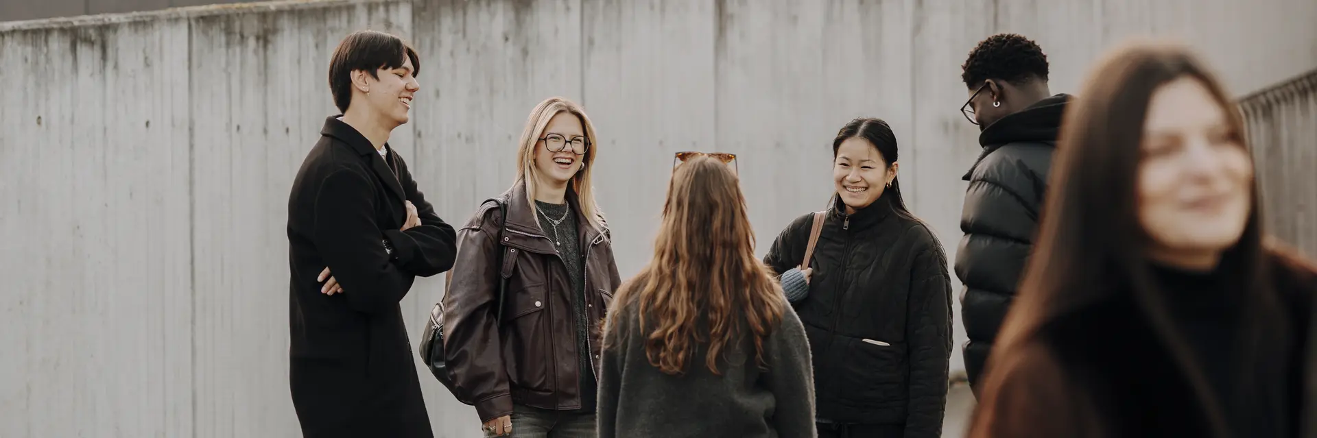Sechs Personen stehen in einer Gruppe und unterhalten sich lebhaft. Sie tragen lässige Kleidung und wirken freundlich. Im Hintergrund sind Gebäude und Wandkunst zu sehen, während einige Personen verstreut im Hintergrund stehen.