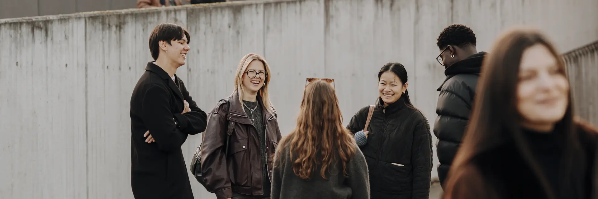 Sechs Personen stehen in einer Gruppe und unterhalten sich lebhaft. Sie tragen lässige Kleidung und wirken freundlich. Im Hintergrund sind Gebäude und Wandkunst zu sehen, während einige Personen verstreut im Hintergrund stehen.