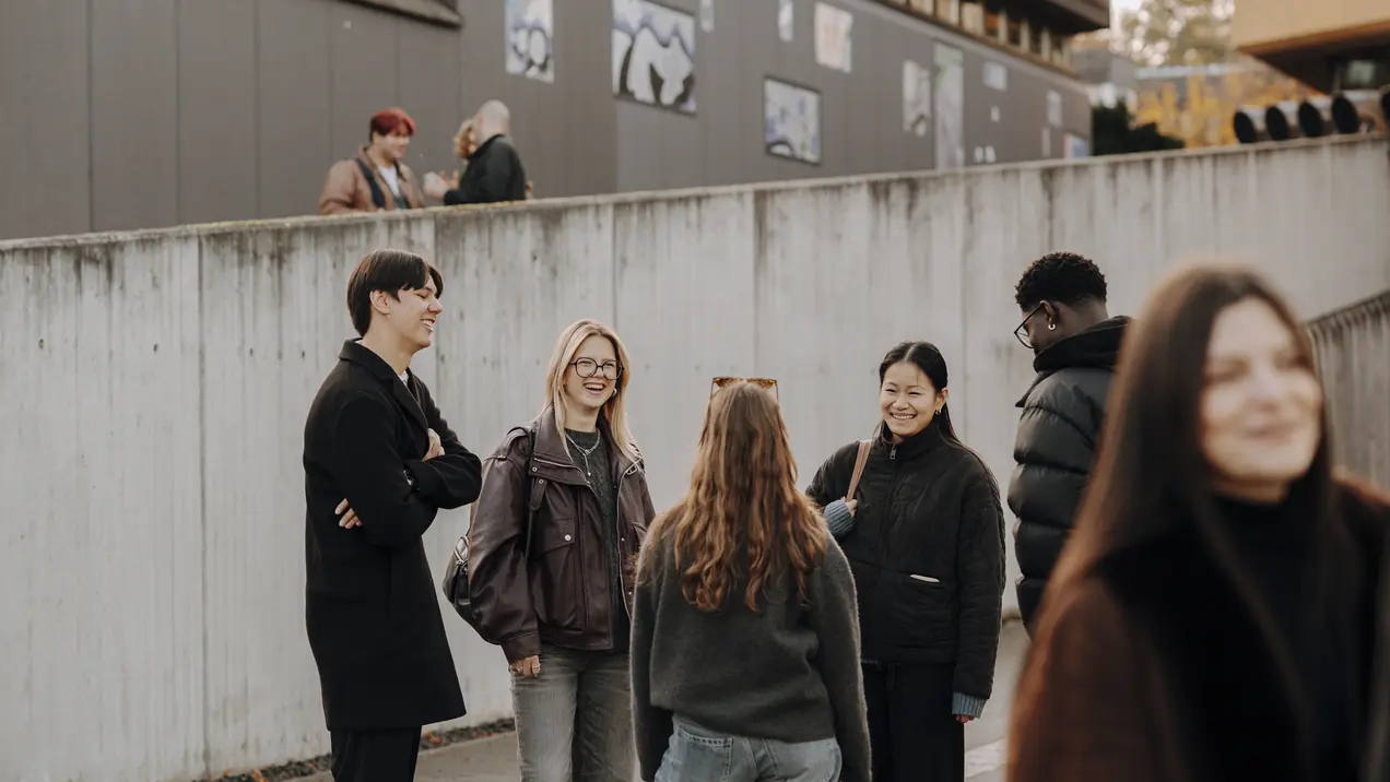 Sechs Personen stehen in einer Gruppe und unterhalten sich lebhaft. Sie tragen lässige Kleidung und wirken freundlich. Im Hintergrund sind Gebäude und Wandkunst zu sehen, während einige Personen verstreut im Hintergrund stehen.