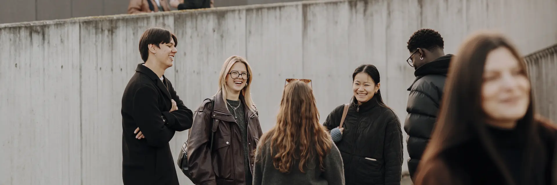Sechs Personen stehen in einer Gruppe und unterhalten sich lebhaft. Sie tragen lässige Kleidung und wirken freundlich. Im Hintergrund sind Gebäude und Wandkunst zu sehen, während einige Personen verstreut im Hintergrund stehen.