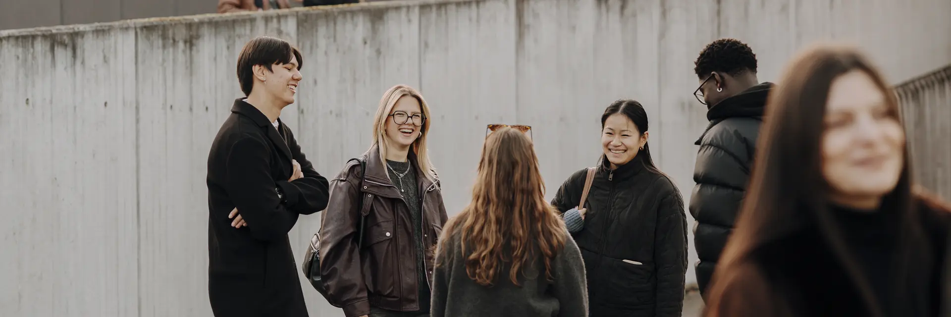 Sechs Personen stehen in einer Gruppe und unterhalten sich lebhaft. Sie tragen lässige Kleidung und wirken freundlich. Im Hintergrund sind Gebäude und Wandkunst zu sehen, während einige Personen verstreut im Hintergrund stehen.