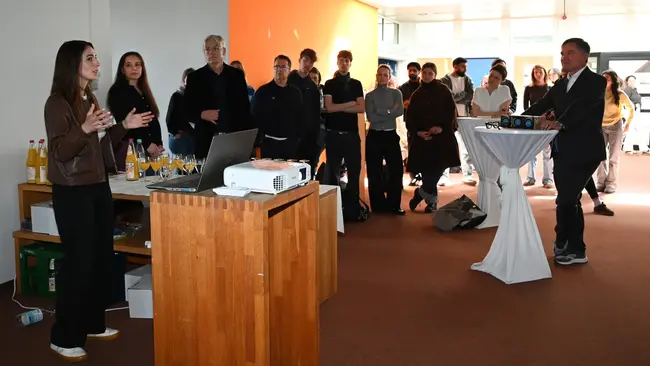 Maral Koohestanian steht an einem Tisch mit einem Laptop und spricht vor einer Gruppe von Zuhörern. Im Hintergrund sind mehrere Personen versammelt, die aufmerksam zuhören. Ein Projektor zeigt möglicherweise Inhalte auf einer Leinwand. Die Szene wirkt informell und interaktiv.