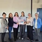 Sechs Frauen stehen in einer Reihe in einem Raum. Sie halten gemeinsam ein Plakat mit dem Text „Gleichstellungsstarke Hochschule“. Einige der Frauen tragen formelle Kleidung, und die Atmosphäre wirkt positiv und kooperativ.