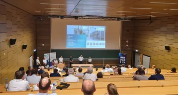 Eine Präsentation in einem Hörsaal, in dem mehrere Personen sitzen. Auf der Bühne stehen fünf Referenten vor einer Leinwand, die Informationen über ein Gebäude zeigt. Der Raum ist gut gefüllt, mit einer modernen Ausstattung und einer Tafel im Hintergrund.