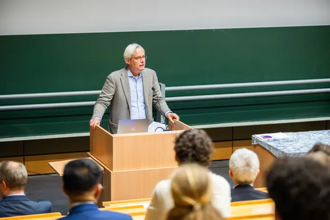 Ein älterer Mann steht an einem Rednerpult in einem auditorischen Raum und hält einen Vortrag. Hinter ihm sitzen Zuhörer, die aufmerksam zuhören. Der Raum ist modern eingerichtet, mit einer grünen Wand und einer Präsentation auf dem Pult.