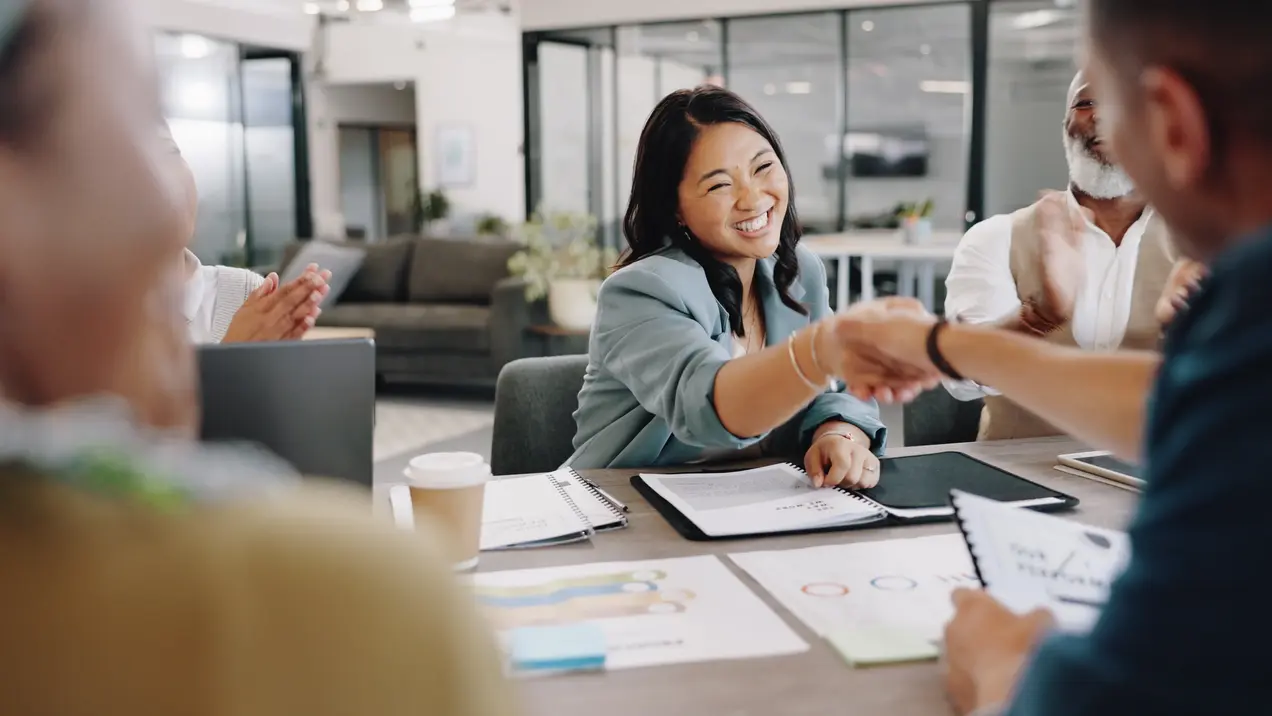 Eine Frau in einem hellblauen Blazer lächelt und schüttelt einer anderen Person die Hand. Im Hintergrund applaudiert eine andere Person. Auf dem Tisch liegen Notizblöcke und Dokumente, während eine entspannte Büroumgebung sichtbar ist.