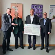 Vier Personen stehen in einem Raum und halten einen großen Scheck über 500 Euro. Die Personen scheinen sich über die Spende zu freuen, während sie in die Kamera schauen. Im Hintergrund sind Plakate und einige Dokumente an der Wand zu sehen.