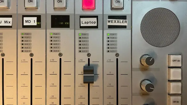 Das Bild zeigt ein Mischpult mit verschiedenen Reglern und Knöpfen. Zu sehen sind die Beschriftungen „J Mix“, „MD 1“, „Laptop“ und „WEXXLER“. Ein roter Knopf ist hervorgehoben. Es gibt mehrere Schieberegler und Kontrollleuchten zur Anpassung der Audiopegel.