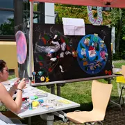 Eine Person sitzt an einem Tisch und arbeitet an Bastelmaterialien. Im Hintergrund ist eine Station mit zwei großen, bunten Plakaten, die fantasievolle Welten darstellen. Über den Plakaten hängt ein Schild mit der Aufschrift „Unseres Tomorrows“. Der Ort ist im Freien, umgeben von Gras und Bäumen.